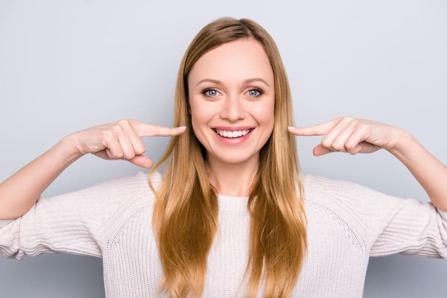 Smiling blonde woman pointing to her professionally whitened teeth.